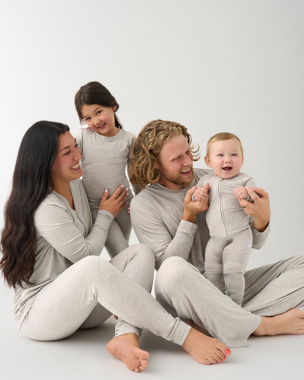 A family of four wearing matching light gray long-sleeve pajamas sits together on a plain light gray studio background, smiling and laughing.