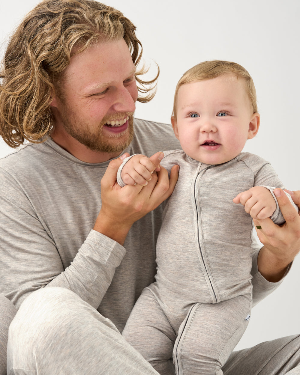 A man and a baby wearing matching light gray heathered pajamas, sitting together against a plain light gray background.