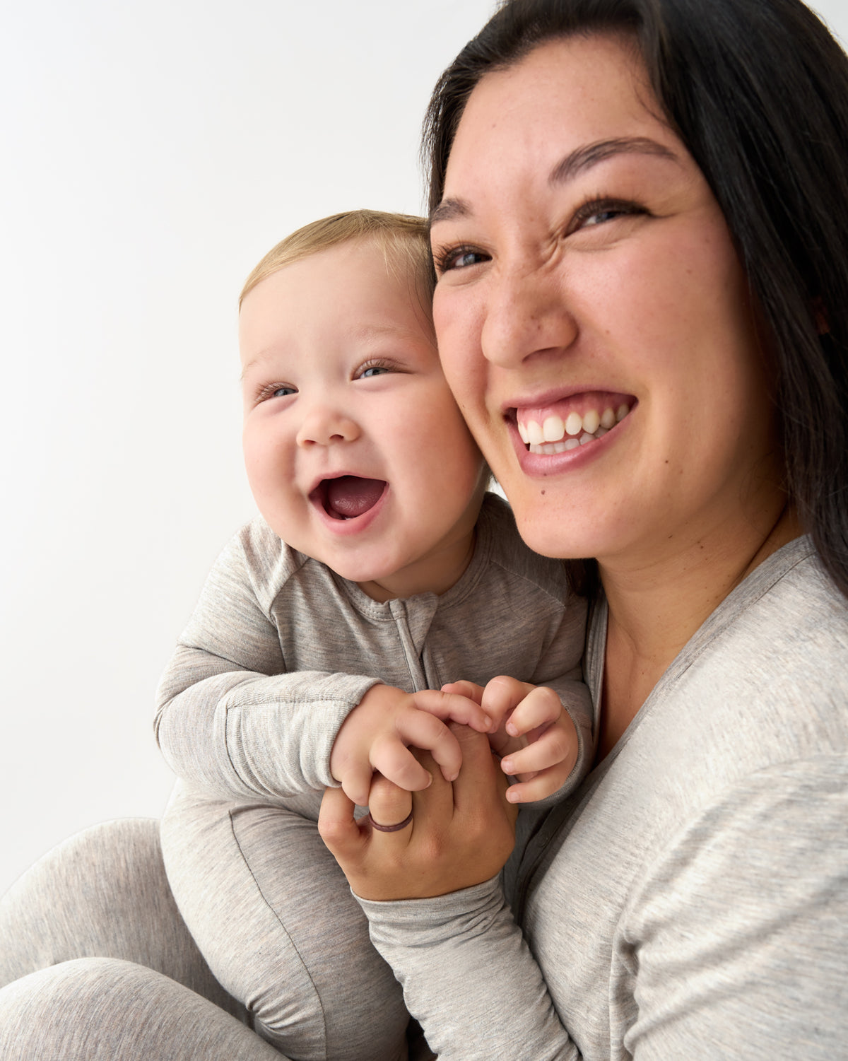 A smiling woman holding a laughing baby, both wearing matching light gray long-sleeve one-piece outfits, against a plain white background.