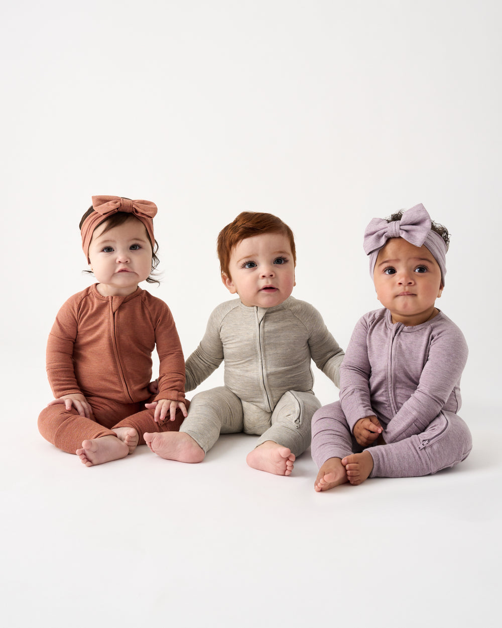 Three babies sitting side by side wearing matching one-piece footless jumpsuits and bow headbands in dusty rose, beige, and lavender on a white studio background.
