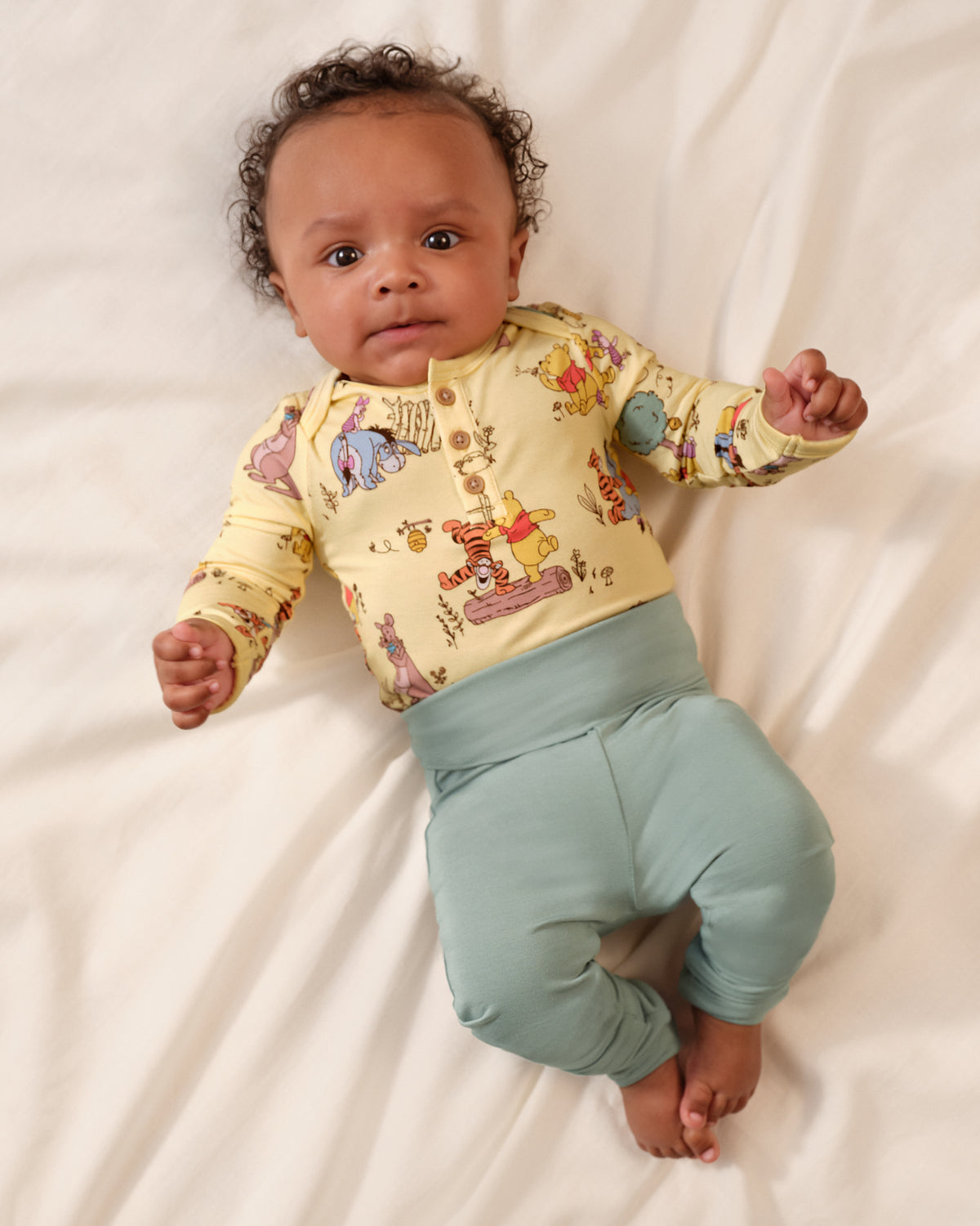 A baby lying on white sheets, wearing a long-sleeve button-up top in pale yellow with a colorful cartoon animal print and light blue-green pants.