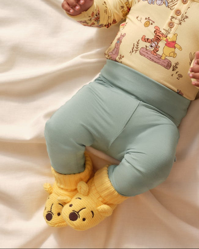 Baby wears a yellow top with a Winnie the Pooh print and sage green leggings with yellow slippers.