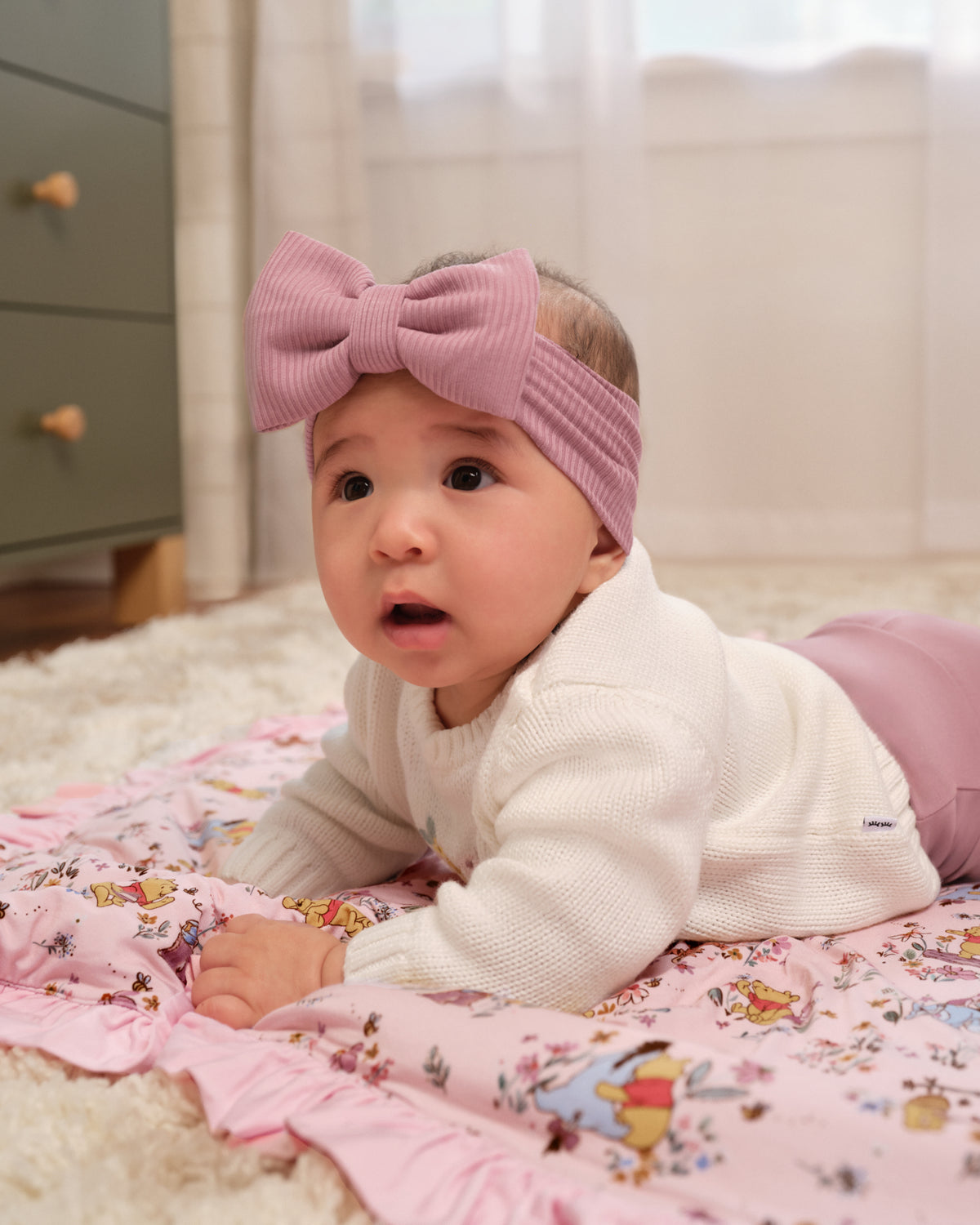 A baby lying on a pink patterned blanket on a white rug, wearing a large pink ribbed bow headband and a white knit top.