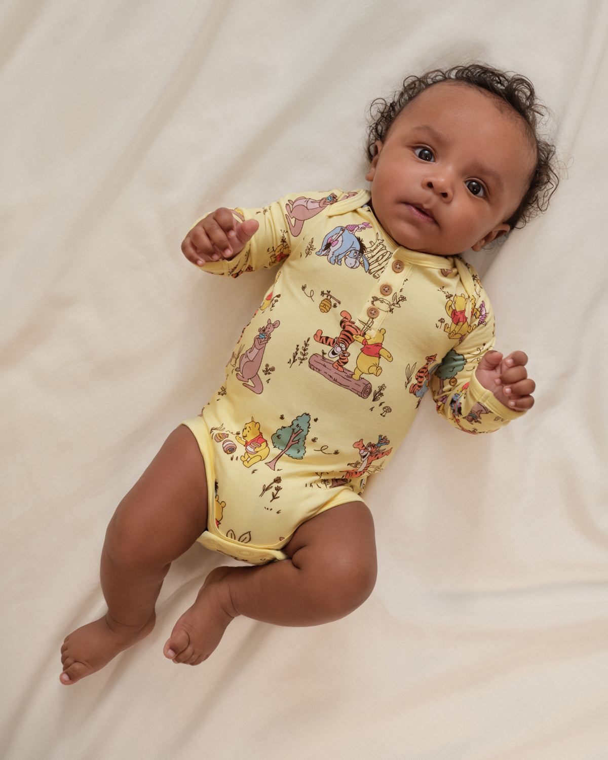 A baby lying on a white fabric surface wearing a pale yellow long-sleeve bodysuit with a colorful cartoon print featuring Winnie the Pooh characters.