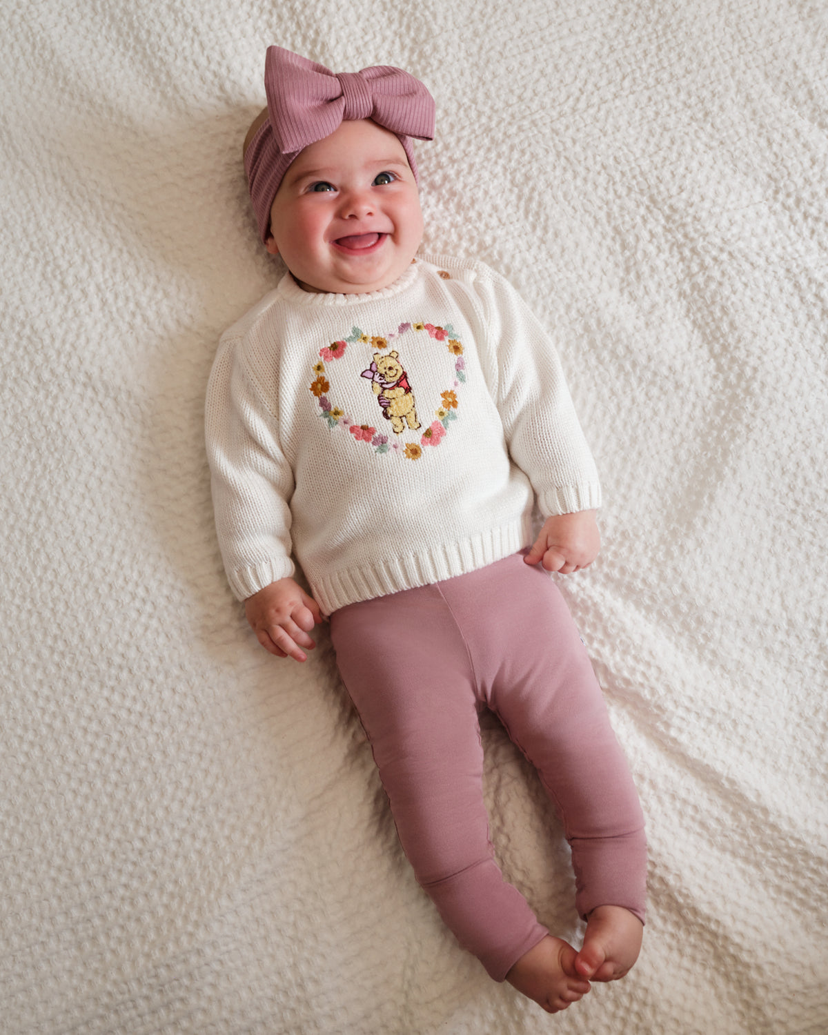 A smiling baby lying on a textured cream blanket, wearing a cream knit sweater with a colorful floral heart design, dusty pink leggings, and a matching dusty pink bow headband.