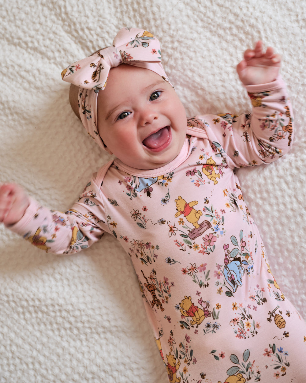 A smiling baby lying on a textured cream blanket, wearing a light pink long-sleeve bodysuit with a multicolor Winnie the Pooh print and a matching pink bow headband.