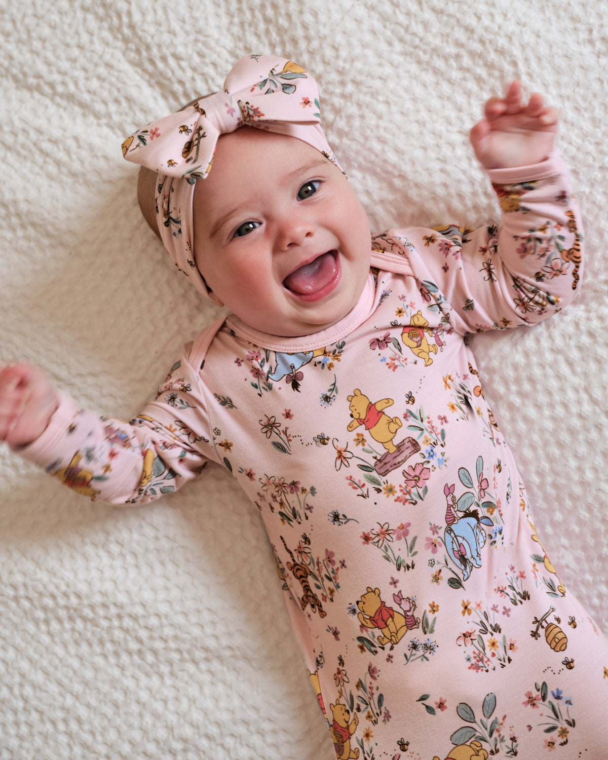A smiling baby lying on a textured cream blanket, wearing a light pink long-sleeve bodysuit with a multicolor Winnie the Pooh print and a matching pink bow headband.