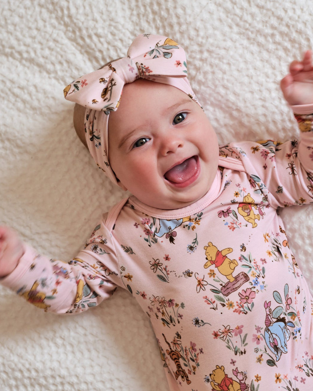 A smiling baby lying on a textured cream blanket, wearing a light pink long-sleeve bodysuit with a multicolor Winnie the Pooh print and a matching pink bow headband.