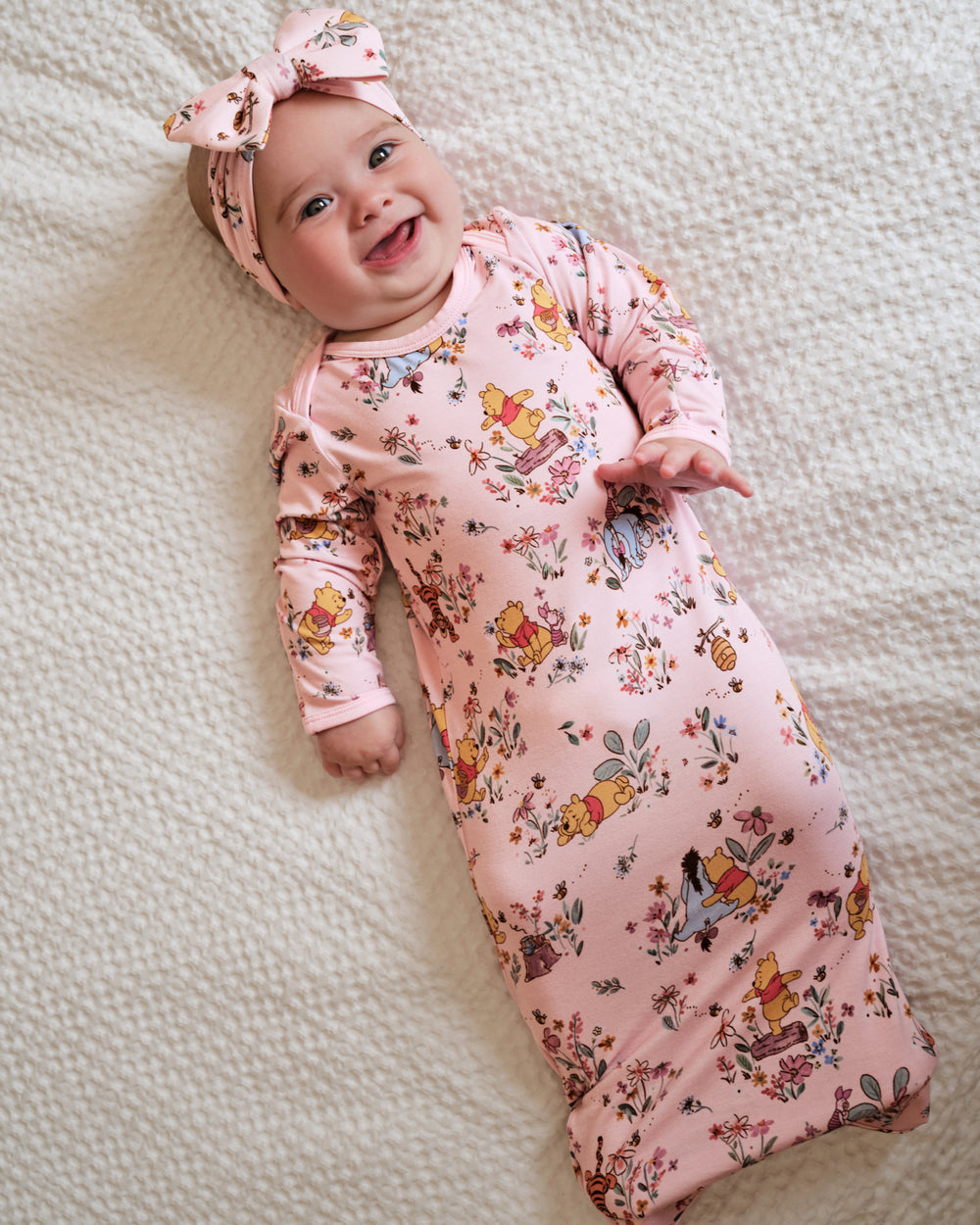 A smiling baby lying on a textured cream blanket, wearing a pink long-sleeve sleep sack and matching bow headband with a colorful Winnie the Pooh print.