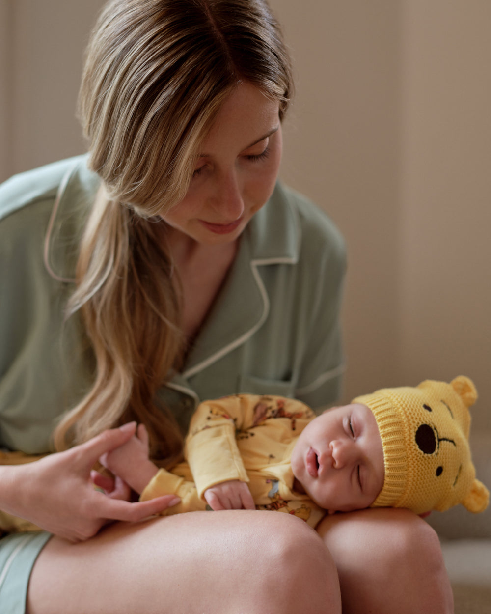 A woman in sage green pajamas holds a sleeping newborn baby wearing a yellow knit onesie and a yellow bear-face bonnet.