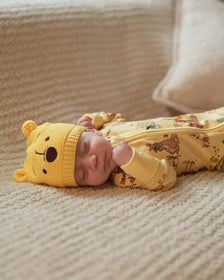 A sleeping newborn baby wearing a yellow knit hat with a bear face and a matching yellow printed footed sleeper, lying on a textured beige blanket.
