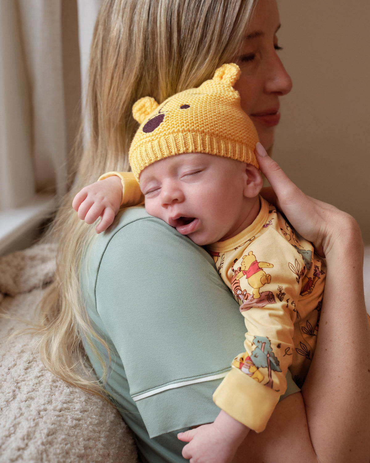 A sleeping newborn baby is cradled in the arms of a woman. The baby is wearing a yellow knit bear hat with a stitched nose and a yellow long-sleeve onesie printed with Winnie the Pooh characters.