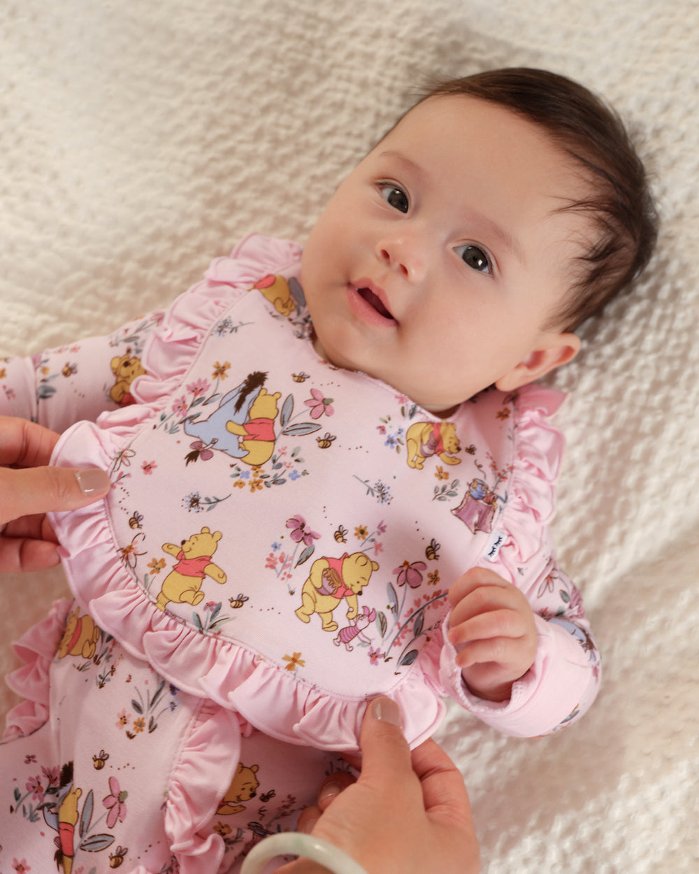 A baby lying on a textured cream blanket, wearing a pink long-sleeve romper with Winnie the Pooh print and ruffled trim. Hands are adjusting the ruffled collar area of the garment.