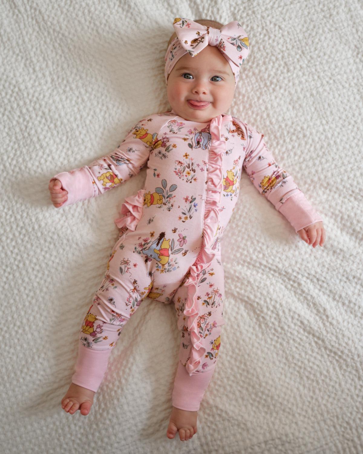 A baby lying on a white textured blanket, wearing a pink long-sleeve footless onesie with a floral and fairy print, ruffle trim, and a matching pink bow headband.