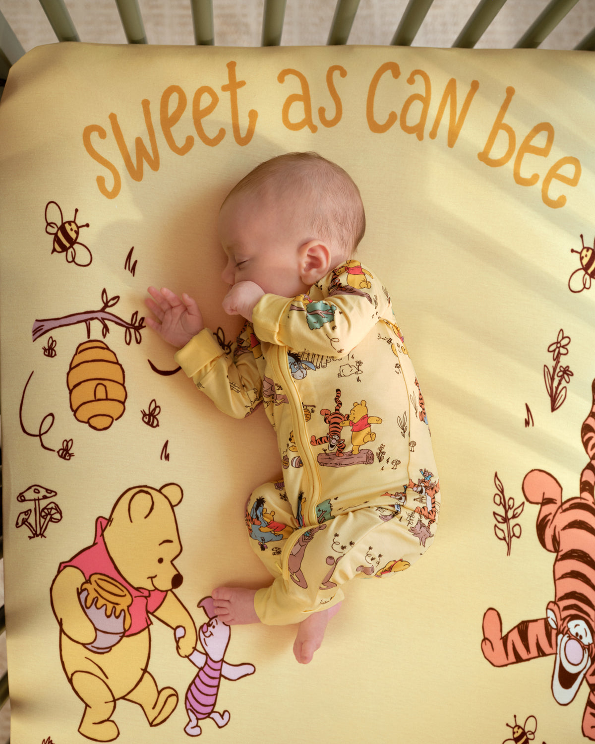 A baby sleeping in a crib wearing a yellow long-sleeve onesie with a colorful cartoon print, lying on a matching yellow crib mattress printed with 'Sweet as can bee' and Winnie the Pooh characters.