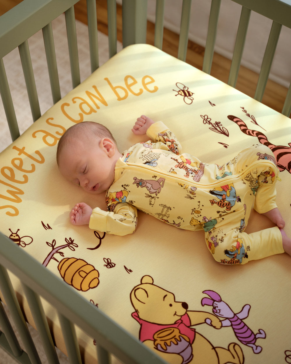 A baby wearing a yellow long-sleeve footed sleeper with a Winnie the Pooh print lies on their side in a crib. The crib mattress has a matching yellow cover with 'sweet as can bee' text and cartoon illustrations.