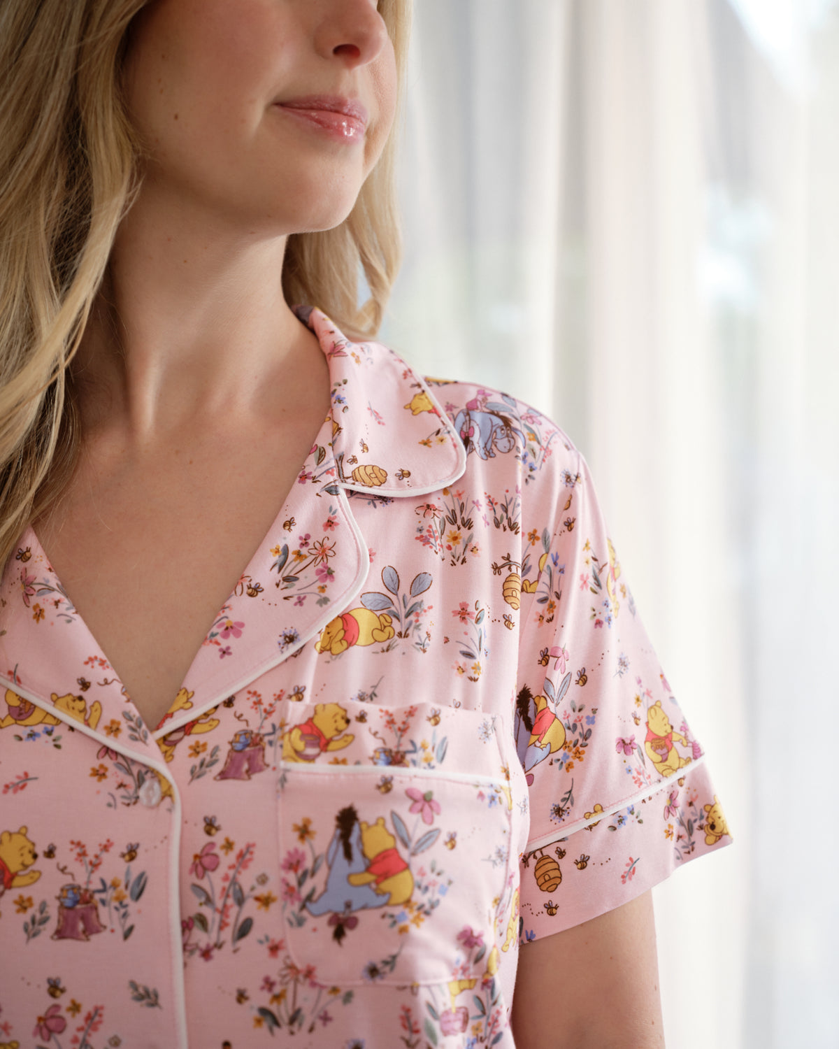 A close-up of a woman wearing a light pink short-sleeve pajama shirt with a Winnie the Pooh floral and character print, featuring a collar and button placket.