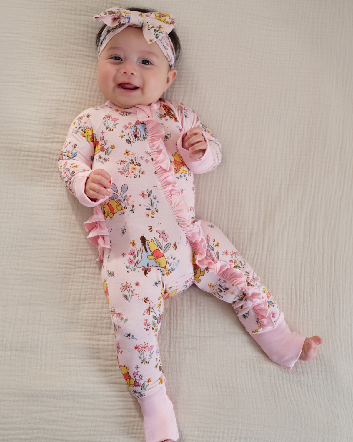 A baby lying on a textured cream blanket, wearing a pink footed sleeper with a floral and bird print, ruffle trim, and a matching floral headband.