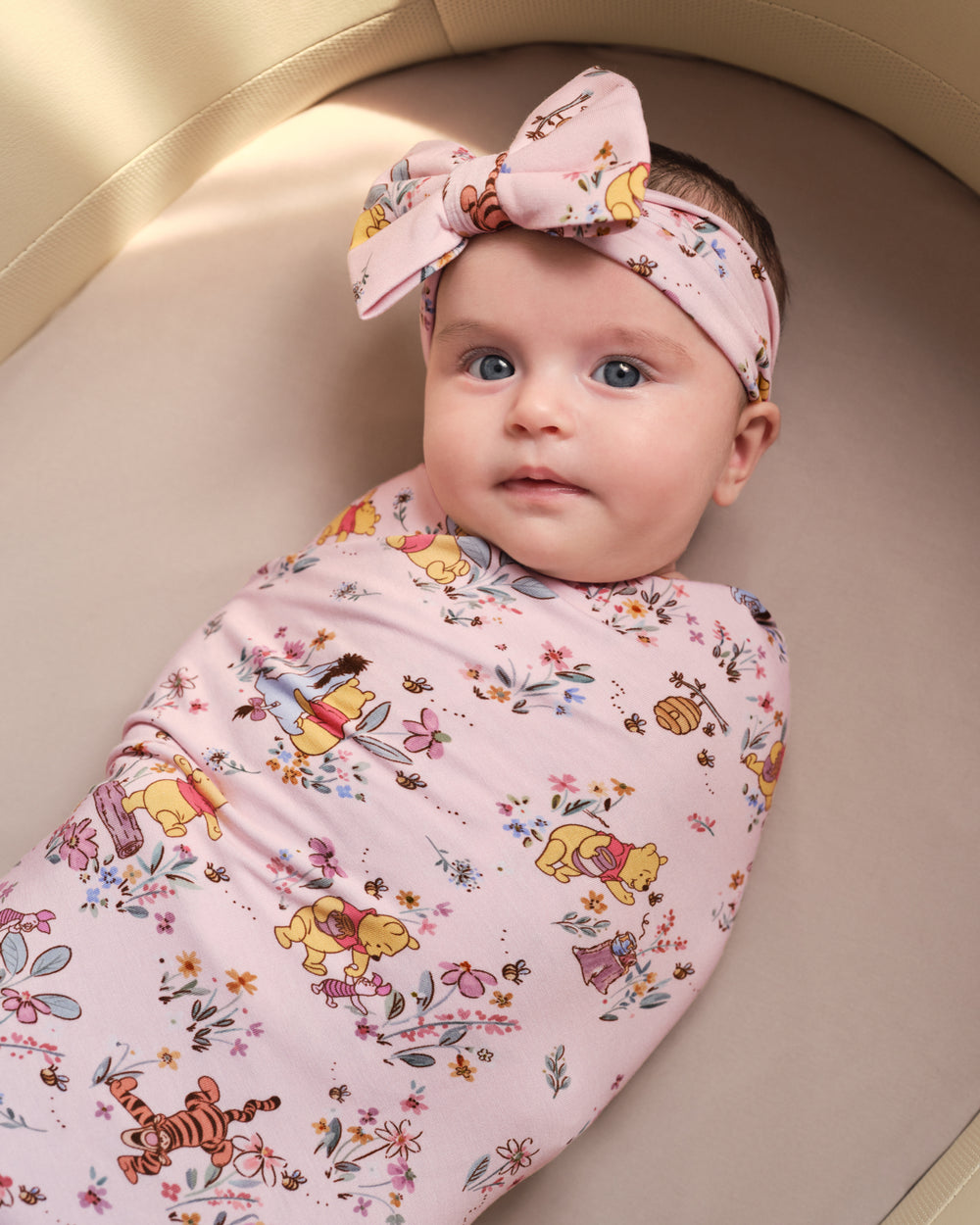 A baby lying on their back inside a beige bassinet, swaddled in a light pink muslin swaddle blanket with a Winnie the Pooh and Tigger print, and wearing a matching pink bow headband.