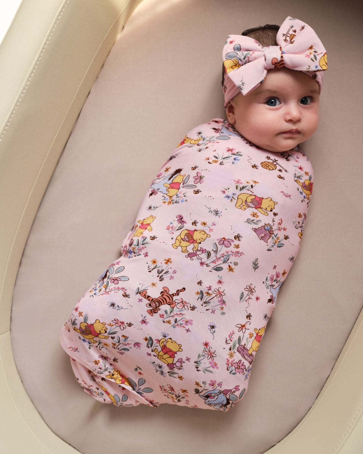 A baby wrapped in a pink floral and cartoon print swaddle blanket, wearing a matching pink bow headband, lying inside a beige bassinet.