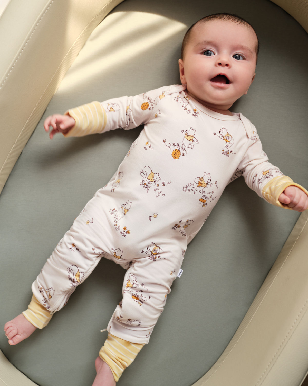 A baby lying on their back inside a bassinet, wearing a cream-colored long-sleeve footless sleeper with a yellow and brown honeybee pattern and yellow striped cuffs.