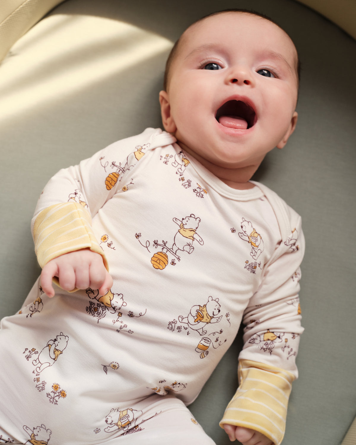 A baby lying on their back wearing a cream-colored long-sleeve footed sleeper with a yellow and brown cartoon bear print and yellow striped cuffs, set against a soft gray cushion.