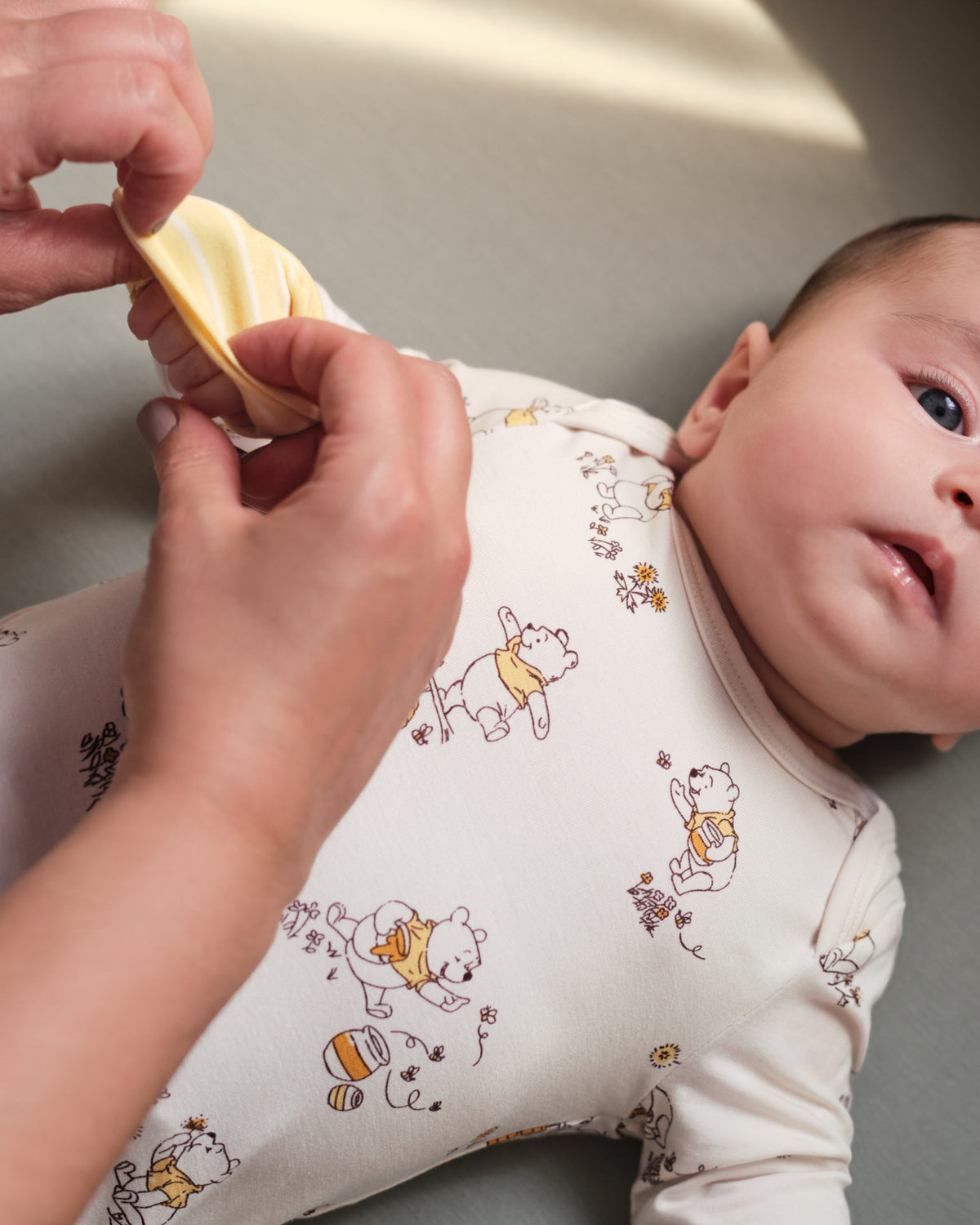 A baby lying on their side wearing a cream-colored long-sleeve bodysuit with a Winnie the Pooh print, while an adult's hands adjust a matching yellow and white bib on the baby's chest.