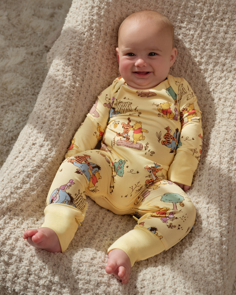 A smiling baby lying on a textured cream blanket, wearing a pale yellow long-sleeve footed sleeper with a colorful Winnie the Pooh print.