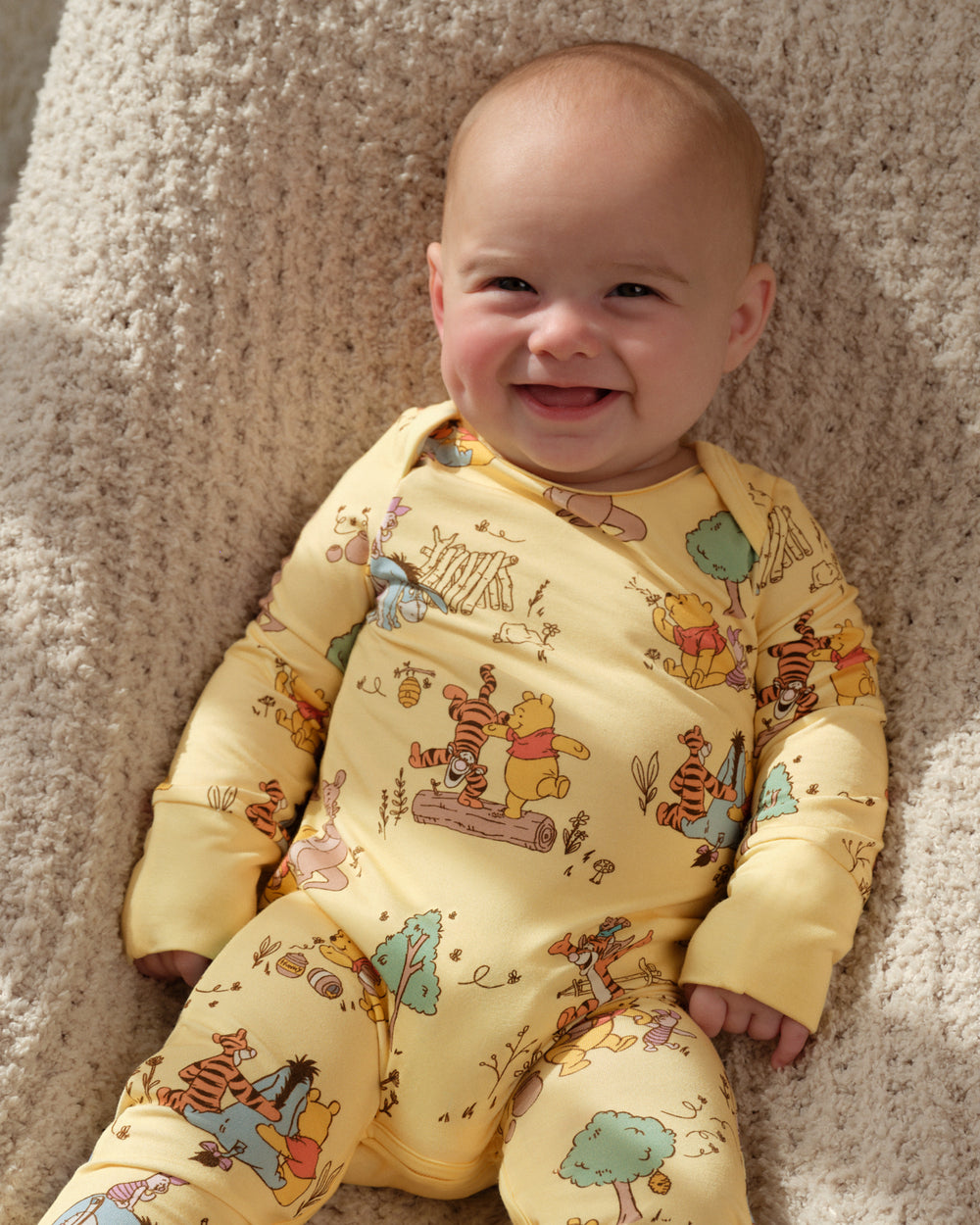 A smiling baby lying on a beige textured blanket, wearing a yellow long-sleeve footed sleeper with a colorful Winnie the Pooh print.