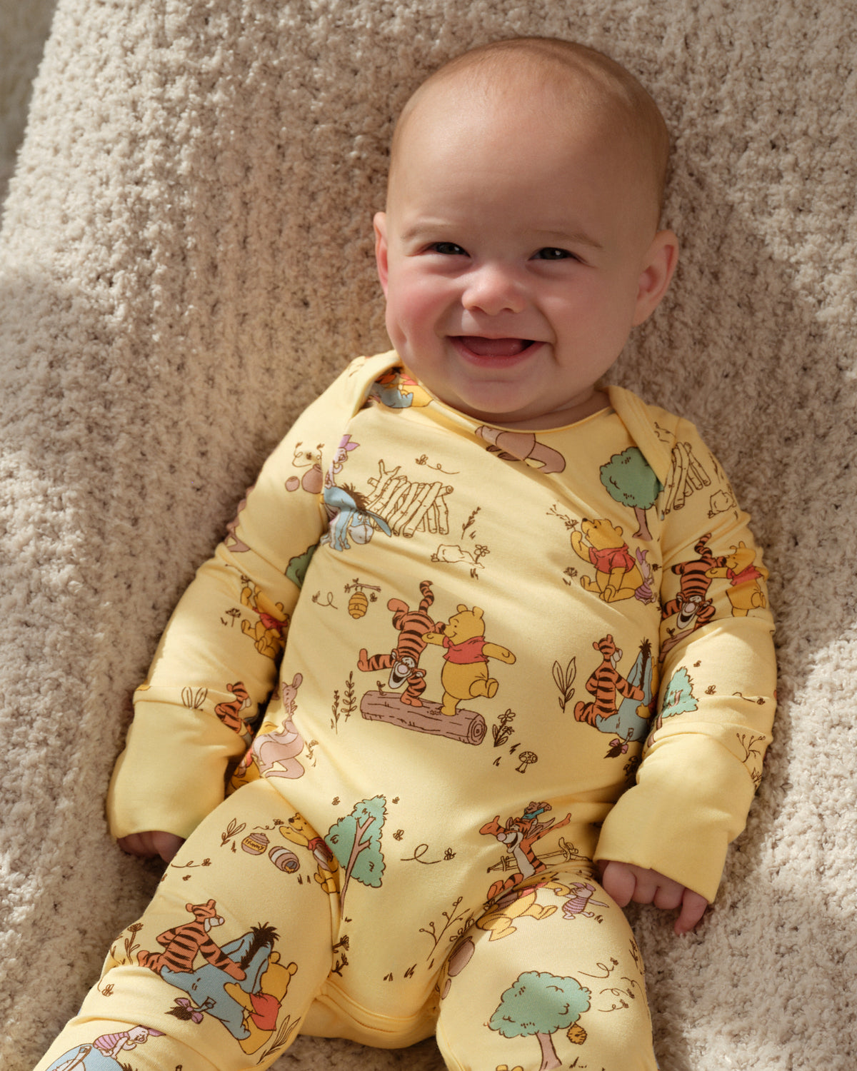 A smiling baby lying on a beige textured blanket, wearing a yellow long-sleeve footed sleeper with a colorful Winnie the Pooh print.