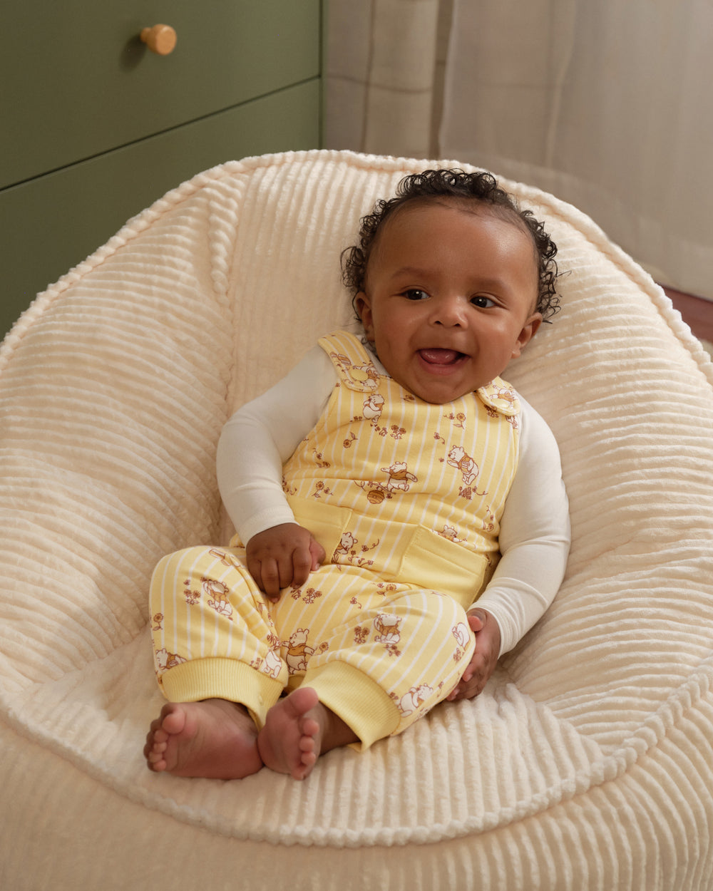 A baby sitting in a cream ribbed beanbag chair, wearing a yellow and white striped sleeveless romper with a teddy bear print over a white long-sleeve shirt.