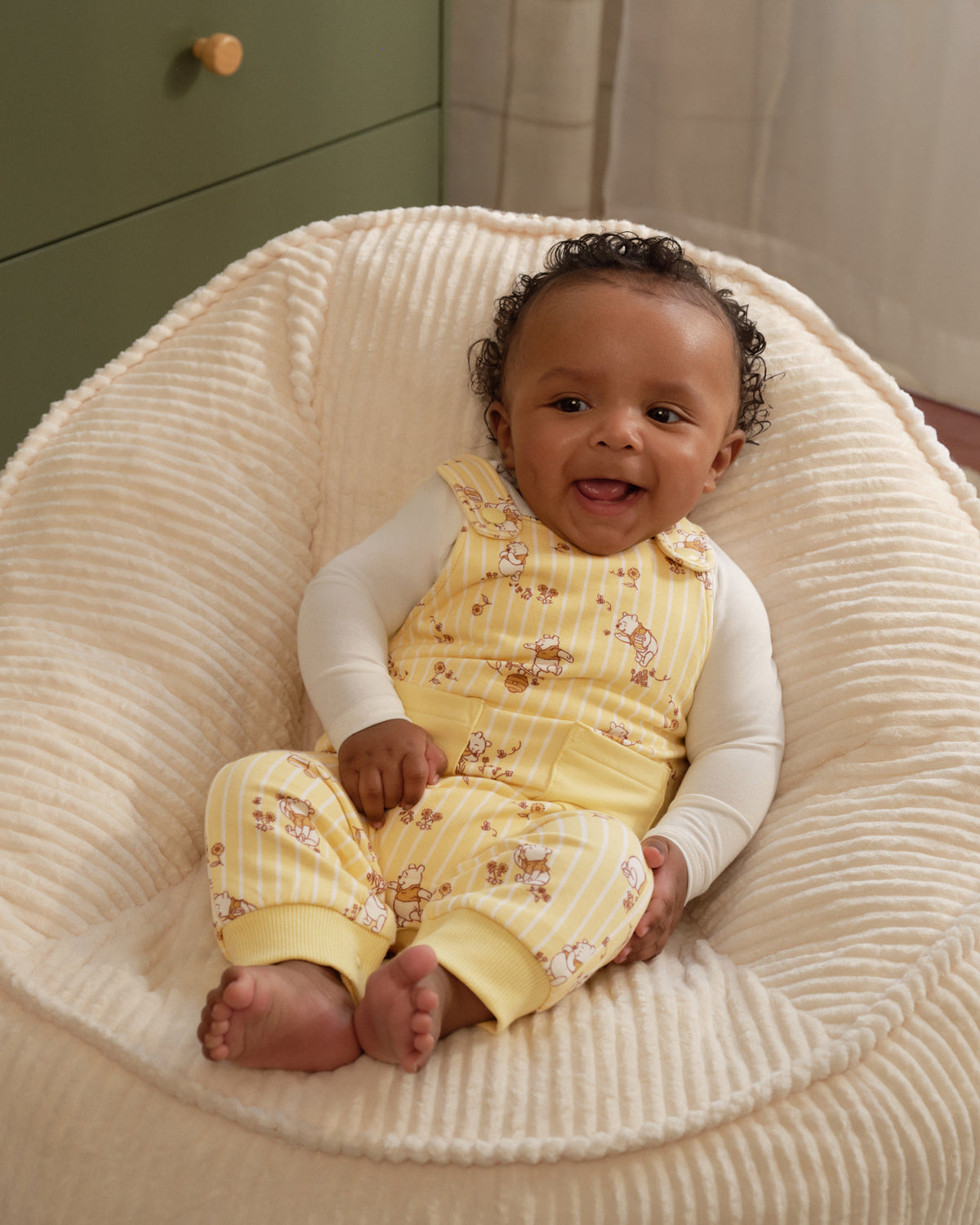 A baby sitting in a cream ribbed beanbag chair, wearing a yellow and white striped sleeveless romper with a teddy bear print over a white long-sleeve shirt.