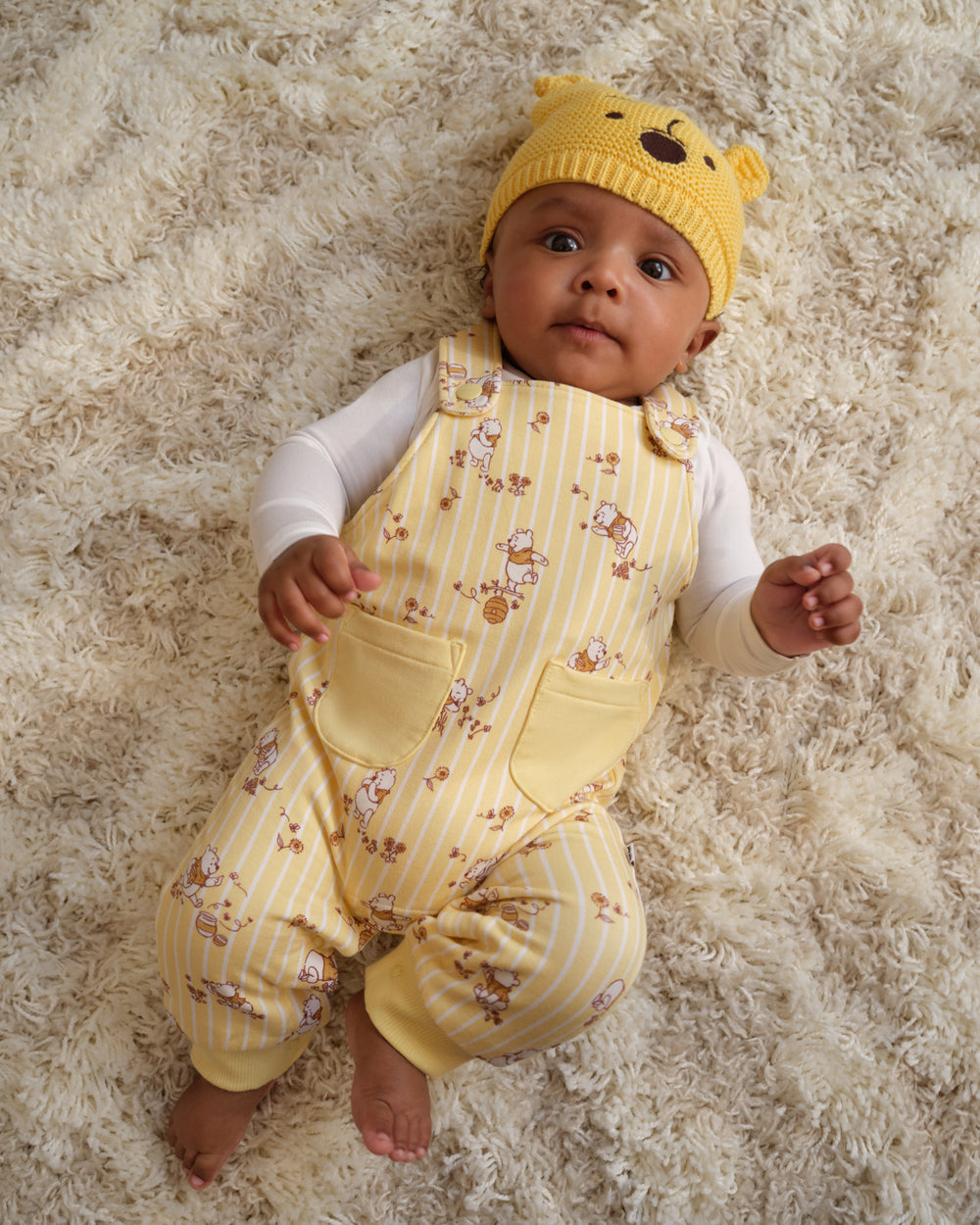 A baby lying on a fluffy cream shag rug, wearing a yellow knit hat with bear ears and a yellow and white striped overall romper with a small teddy bear print and white long-sleeve shirt.