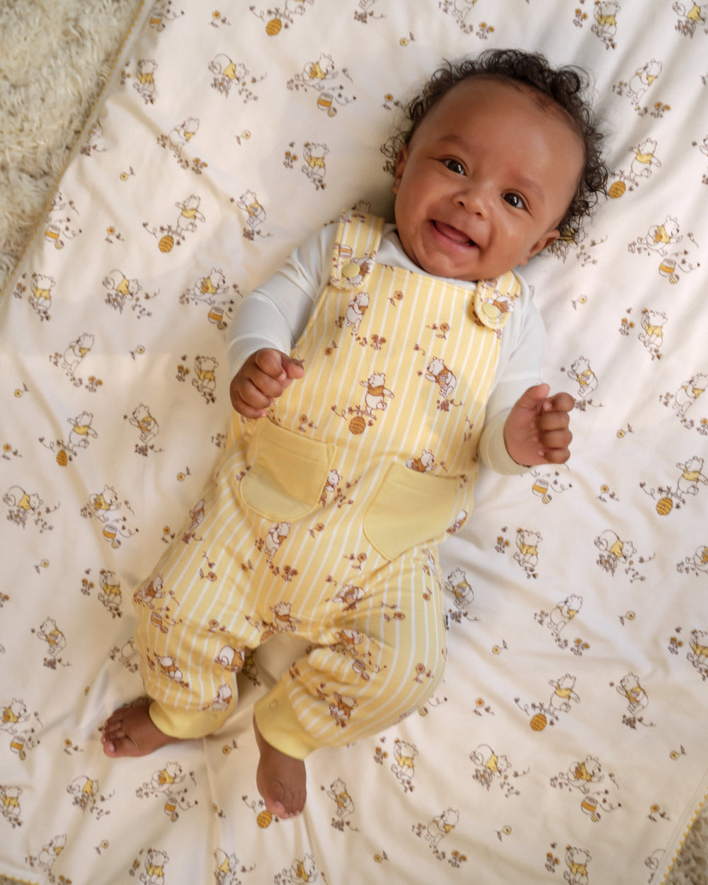 A smiling baby lying on a patterned cream blanket, wearing pale yellow and white striped overalls with a white long-sleeve shirt.