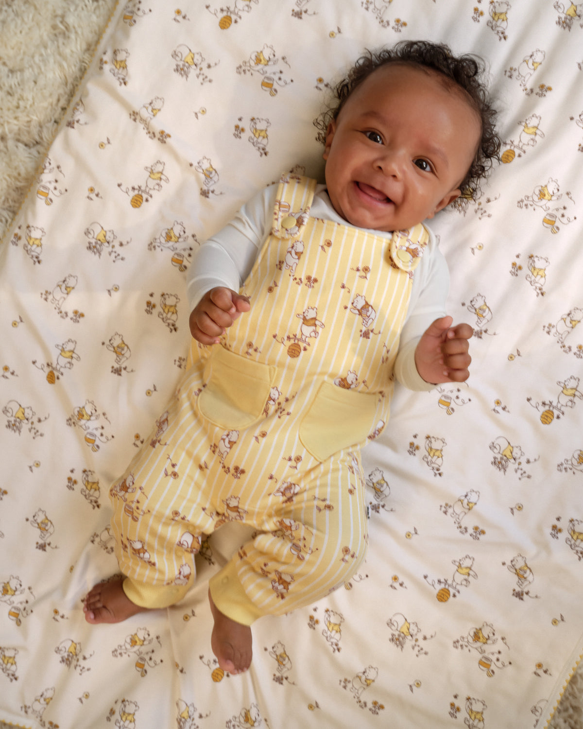 A smiling baby lying on a patterned cream blanket, wearing pale yellow and white striped overalls with a white long-sleeve shirt.