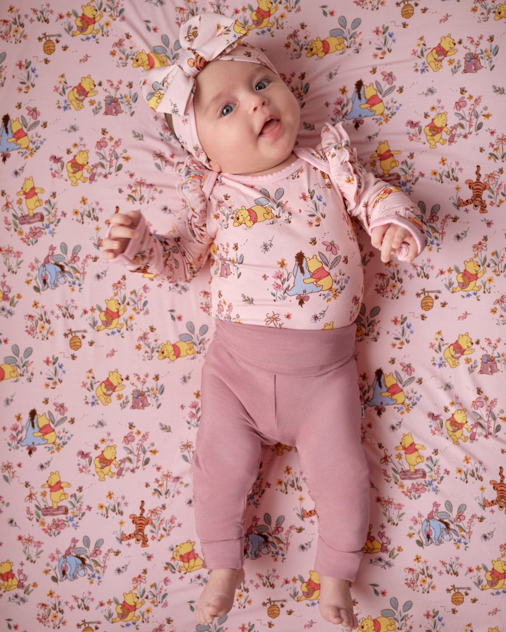 A baby lying on a pink floral and cartoon-patterned blanket, wearing a matching pink long-sleeve top, pink leggings, and a pink bow headband.