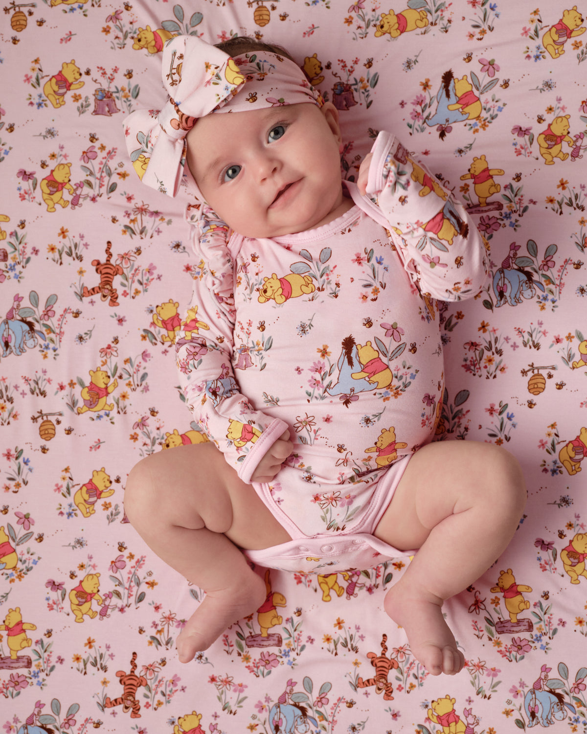 A baby lying on a pink Winnie the Pooh print blanket, wearing a matching pink long-sleeve bodysuit and bow headband with a Winnie the Pooh pattern.
