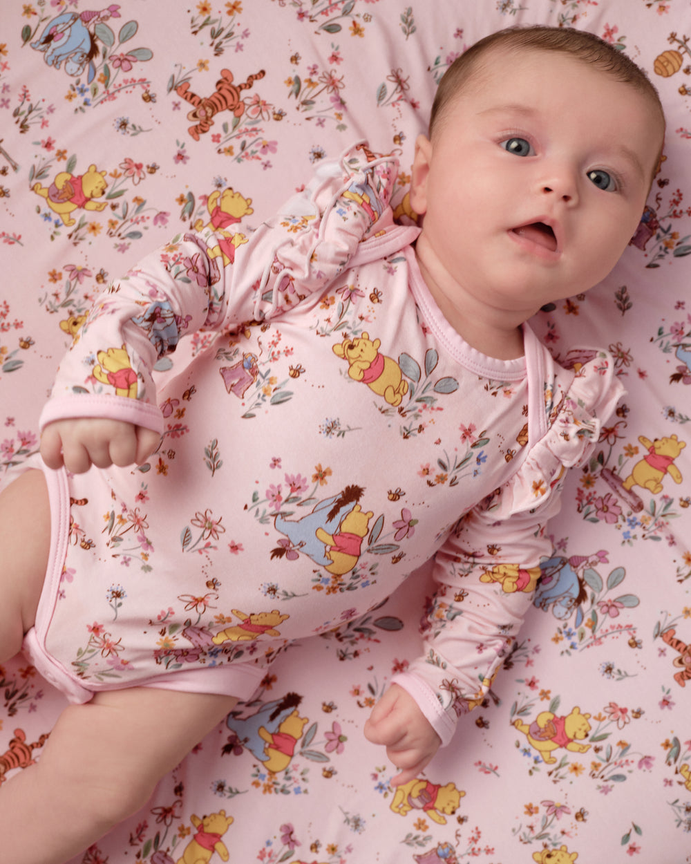 A baby lying on a pink floral and cartoon print sheet, wearing a matching long-sleeve pink bodysuit with Winnie the Pooh and other character prints.