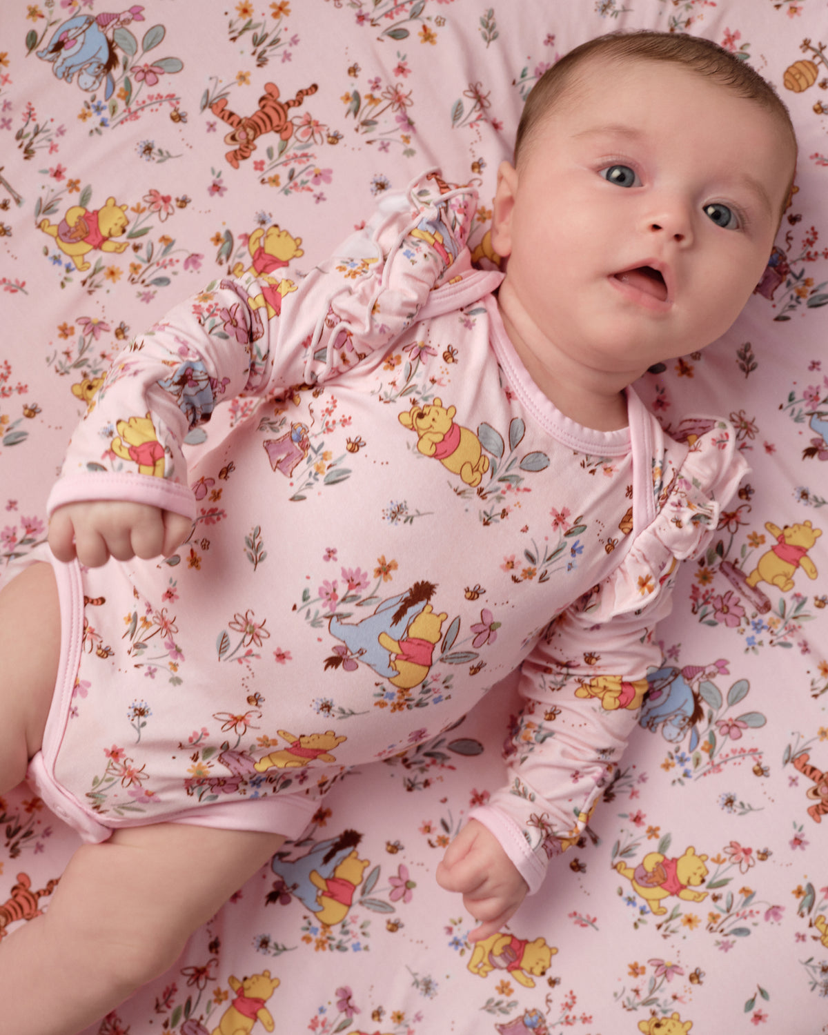 A baby lying on a pink floral and cartoon print sheet, wearing a matching long-sleeve pink bodysuit with Winnie the Pooh and other character prints.