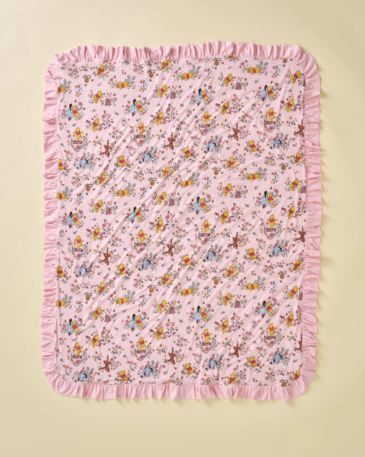 A square pink quilted blanket with a delicate multicolor floral and fairy pattern, finished with a soft pink ruffled border, laid flat on a light beige background.
