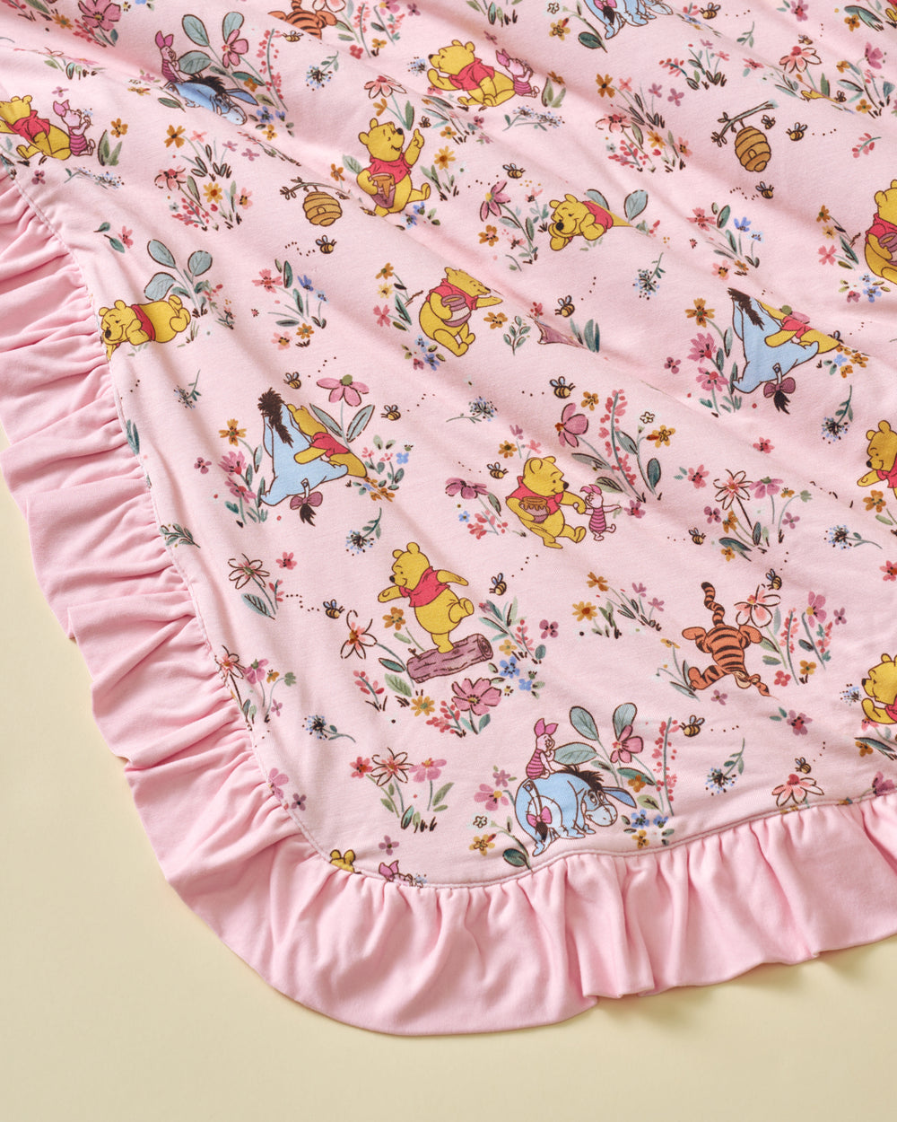 A pink blanket featuring a repeating pattern of Winnie the Pooh, Tigger, Eeyore, and other beloved cartoon characters surrounded by small flowers, finished with a soft pink ruffled border.