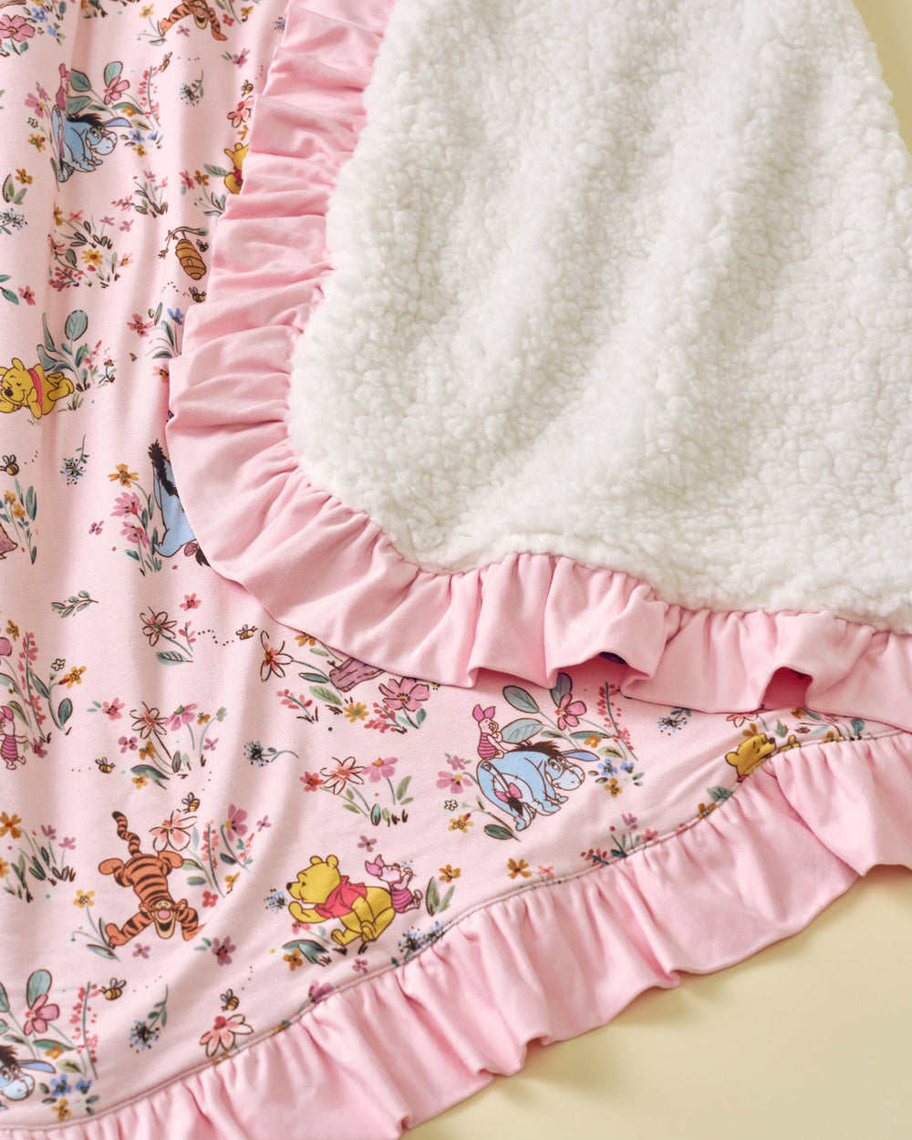 Close-up of a pink ruffled fabric garment or bedding piece featuring a Winnie the Pooh and Tigger floral print, with a white sherpa fleece backing visible along the ruffle edge.