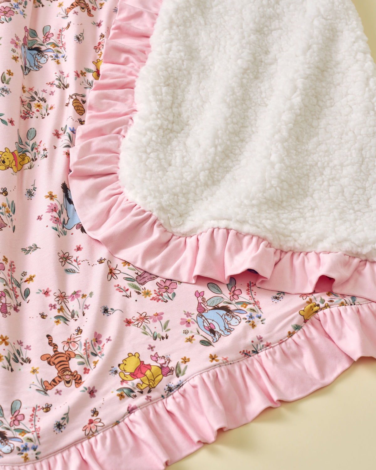 Close-up of a pink ruffled fabric garment or bedding piece featuring a Winnie the Pooh and Tigger floral print, with a white sherpa fleece backing visible along the ruffle edge.