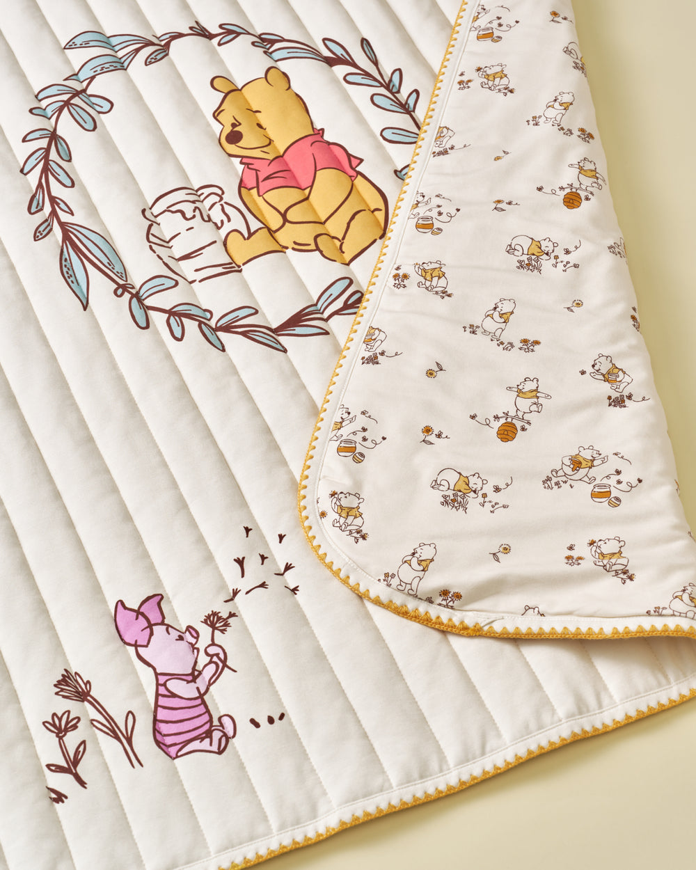 A close-up of a quilted baby blanket featuring Winnie the Pooh and Piglet illustrations. The blanket has a cream-colored quilted side with brown and pink character prints, and a reverse side with a small repeating character pattern on a white background, finished with a yellow zigzag border.