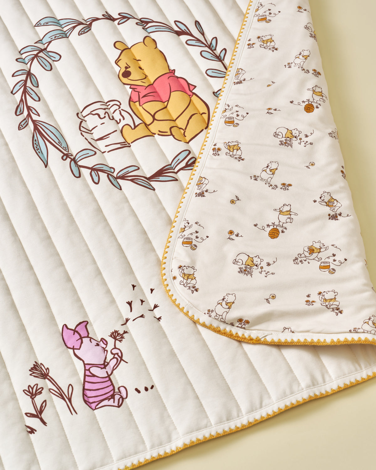 A close-up of a quilted baby blanket featuring Winnie the Pooh and Piglet illustrations. The blanket has a cream-colored quilted side with brown and pink character prints, and a reverse side with a small repeating character pattern on a white background, finished with a yellow zigzag border.
