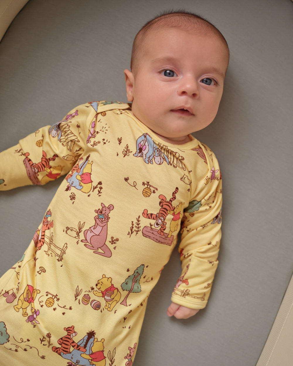 A baby lying on their back wearing a long-sleeve yellow footmuff with a colorful cartoon print of Winnie the Pooh and other Disney characters, set against a neutral gray background.