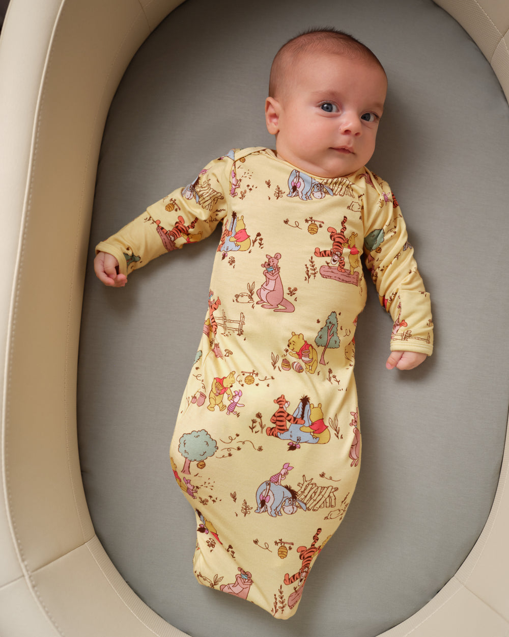A baby lying on their back inside a beige bassinet, wearing a long-sleeve sleep sack with a colorful Winnie the Pooh print on a pale yellow background.