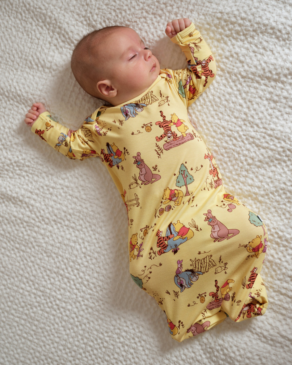 A baby lying on a textured cream blanket, wearing a yellow long-sleeve footless sleeper with a colorful cartoon animal print.