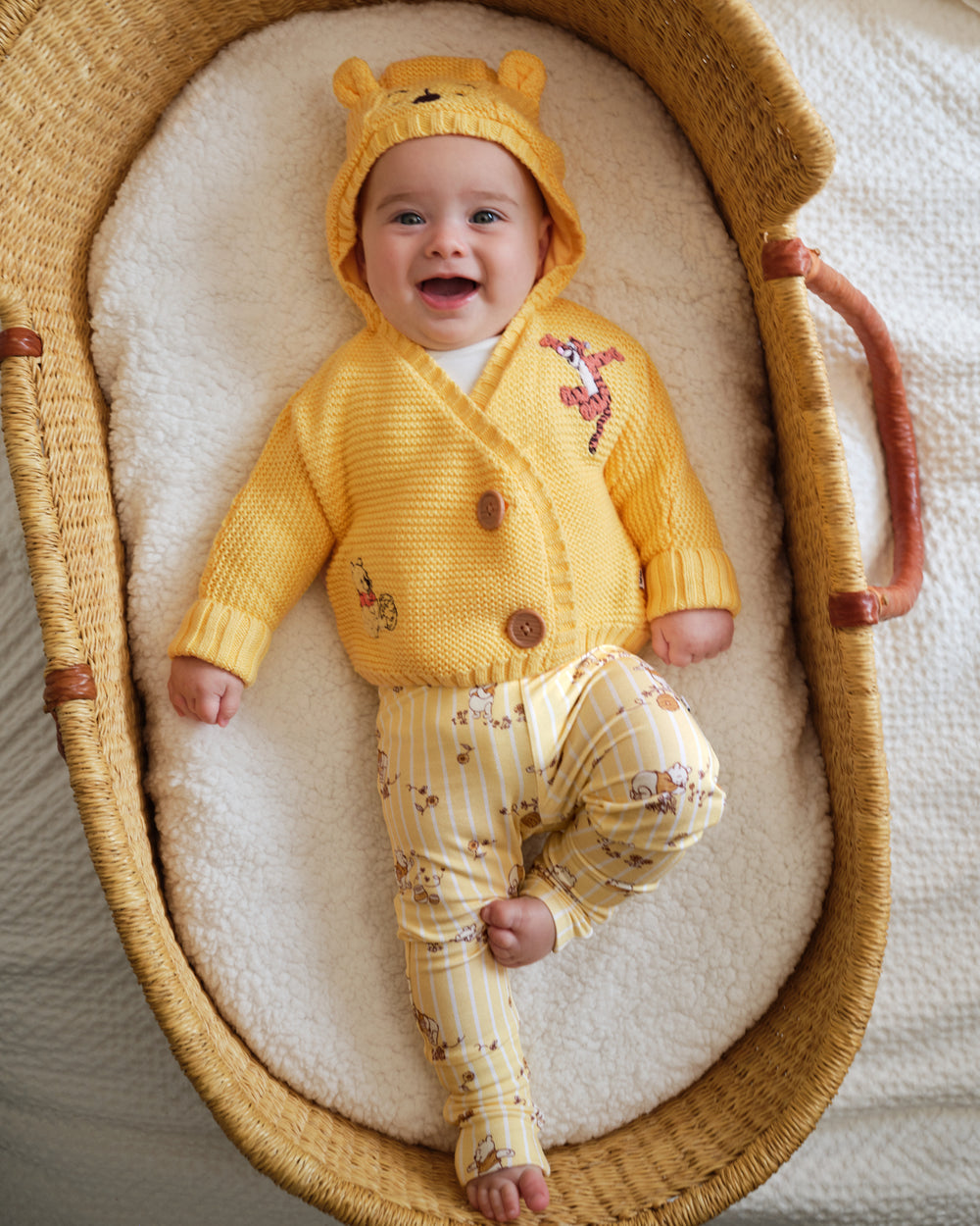 A baby wearing a yellow knit hooded cardigan and matching striped pants with floral prints, lying on a white fleece mattress inside a woven Moses basket.