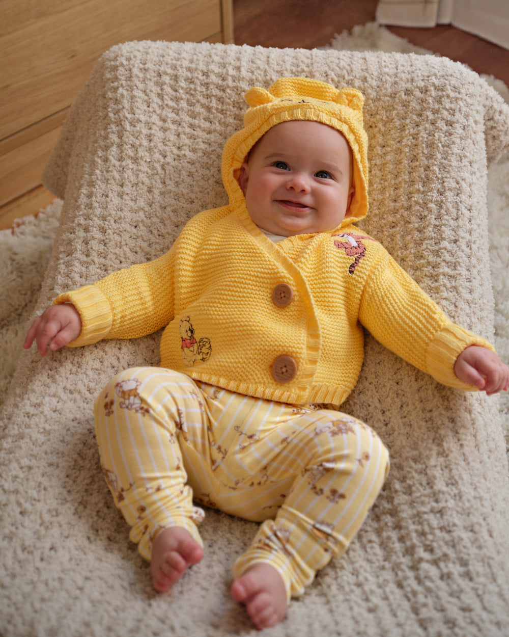 A baby wearing a yellow knit hooded cardigan and matching striped pants with small floral prints, sitting on a textured cream blanket in a home setting.